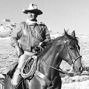 May include: A black and white photo of a man wearing a cowboy hat and leather jacket riding a horse. The man is looking at the camera and the horse is looking to the right. The photo is taken outdoors in a field.