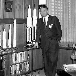 May include: Black and white photograph of a man in a suit standing next to a display of model rockets. The rockets are various sizes and designs, with the text "S-II" visible on one. Bookshelves filled with books are below the rockets.