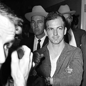May include: A black and white photo of a man in handcuffs being escorted by two men in suits and hats. The man in handcuffs is looking at the camera. The photo has a watermark that says "Do Not Copy. Watermark WILL NOT be on purchased print."