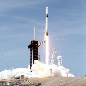 May include: A white and black rocket launches from a launchpad with a large plume of smoke and fire. The rocket is ascending into a blue sky with white clouds.