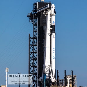 May include: A white and black rocket on a launchpad with the text "SpaceX" on the side. The rocket is attached to a tall metal structure with a platform at the top.