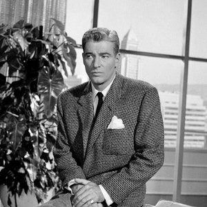 May include: Black and white photograph of a man in a patterned blazer, white shirt, and tie. He is seated with his hands clasped, a pocket square visible. The background includes a large window and a potted plant.