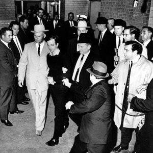 May include: A black and white photo of a group of men in suits. One man is being escorted by two police officers. The man being escorted is wearing a dark shirt and a tie. The police officers are wearing suits and hats. The photo is taken in a hallway with a brick wall in the background.