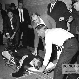 May include: A black and white photo of a man lying on a stretcher, surrounded by people. The man appears to be unconscious. The people are looking down at him with concerned expressions.