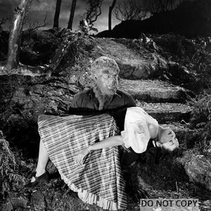 May include: A black and white still from a classic horror film. A werewolf, with a hairy face and chest, cradles a woman in a plaid skirt and white blouse. The scene is set in a dark, wooded area.