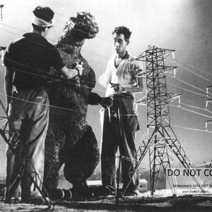 May include: A black and white photo of a man in a dark shirt and a man in a light shirt standing next to a large, gray, dinosaur-like creature. The creature is standing in front of a set of power lines.