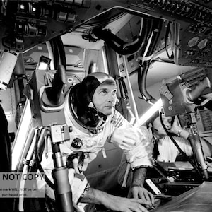 May include: A black and white photo of an astronaut in a spacesuit, sitting in a spacecraft. The astronaut is wearing a helmet and is looking at a control panel. The spacecraft is filled with wires and buttons.