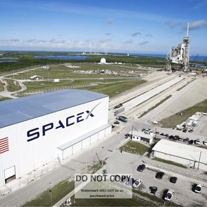 May include: An aerial view of a SpaceX launch site with a large white building labeled "SPACEX" and an American flag on the side. A rocket is visible in the background on a launchpad.