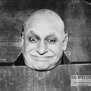 May include: Black and white image of a bald man with a wide grin, peering through a wooden structure. The man's eyes are wide, and he has a prominent nose. The image includes the text "DO NOT COPY" and a watermark.