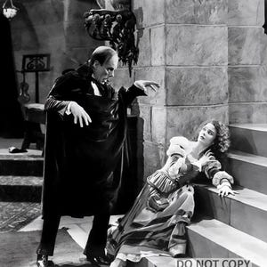 May include: Black and white still from a classic horror film. A man in a dark cloak gestures towards a woman in a period dress, who is seated on stone steps. The scene is set in a gothic interior.