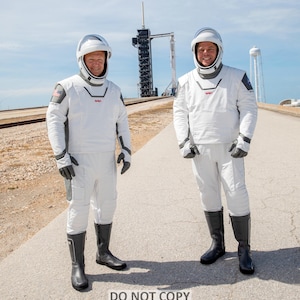 May include: Two astronauts in white spacesuits stand on a paved road in front of a rocket launchpad. The astronauts are wearing helmets and have American flags on their suits. The rocket launchpad is in the background.