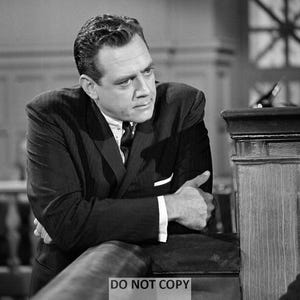 May include: A black and white photo of a man in a suit, looking to the right, with his arms crossed. He is standing in front of a wooden podium.