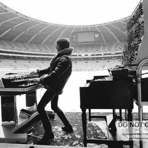 May include: A black and white image of a person playing a keyboard and a piano in a stadium. The person is wearing a jacket and pants. The stadium has a large roof and appears to be empty. There is snow on the ground.