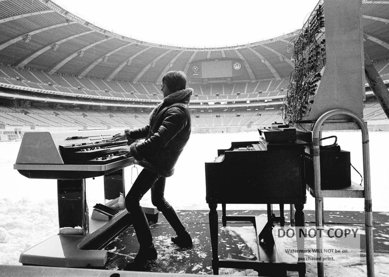 May include: A black and white image of a person playing a keyboard and a piano in a stadium. The person is wearing a jacket and pants. The stadium has a large roof and appears to be empty. There is snow on the ground.