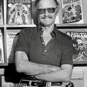 May include: Black and white photograph of a man with glasses and a mustache, arms crossed, smiling. Comic books are visible in the background, including "The Incredible Hulk" and "Marvel's Greatest Superhero Battles by Stan Lee." The image has a watermark that reads "DO NOT COPY."