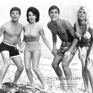 May include: Black and white photo of four people on a beach. Two men and two women are wearing swimsuits. The men are wearing swim trunks and the women are wearing bikinis. The people are smiling and looking at the camera.