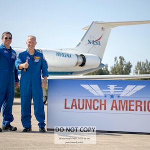 May include: Two astronauts in blue space suits stand in front of a white plane with the word "NASA" on the tail. The plane is parked in front of a sign that reads "LAUNCH AMERICA".