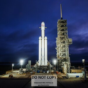 May include: A white rocket with the words "SpaceX" on the side is on a launchpad. The rocket is surrounded by a metal structure and lights. The sky is dark blue with clouds.
