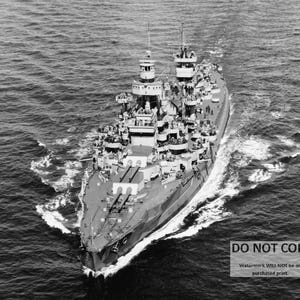 May include: A black and white photograph of a large battleship sailing on the ocean. The ship has multiple gun turrets and is surrounded by white water waves.
