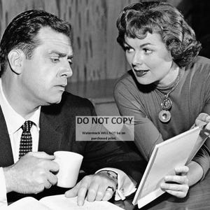 May include: A black and white photo of a man and a woman sitting at a table. The man is wearing a suit and tie, and the woman is wearing a dress. The woman is holding a notebook and a pen. The man is holding a cup.