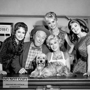 May include: A black and white photo of five people, including two women, two men, and a dog. The women are wearing dresses and the men are wearing shirts and pants. The dog is sitting on a table in front of the people. The photo is taken in a room with a sign that says "Welcome".