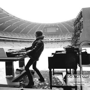 May include: A black and white photograph of a person playing a keyboard and a piano in an empty stadium. The person is wearing a jacket and black pants. A large electronic panel is behind the piano. The stadium is covered in snow.