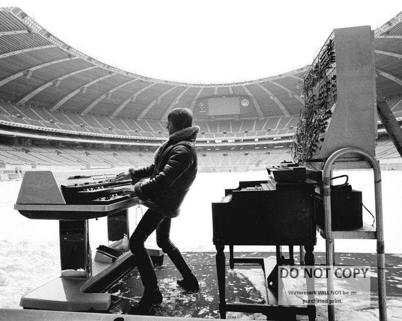 May include: A black and white photograph of a person playing a keyboard and a piano in an empty stadium. The person is wearing a jacket and black pants. A large electronic panel is behind the piano. The stadium is covered in snow.