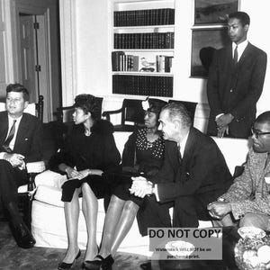 May include: A black and white photo of a group of people sitting in a room. There are three women and three men. The women are wearing dresses and the men are wearing suits. The people are sitting on chairs and a couch. There are bookshelves in the background.