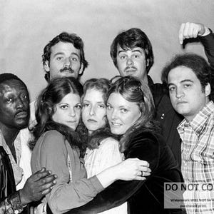 May include: Black and white photograph of a group of seven people, posing closely together. The group includes four women and three men, all with serious expressions. The image is a vintage portrait, likely from the 1970s, with a watermark in the bottom right corner.
