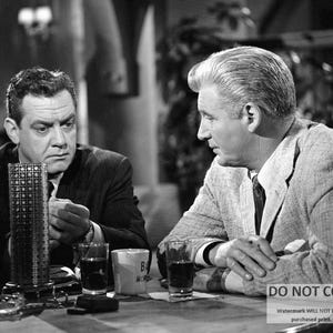May include: Two men in a black and white photo are sitting at a table. The man on the left is wearing a suit and tie. The man on the right is wearing a suit jacket and a tie. They are both looking at the camera.