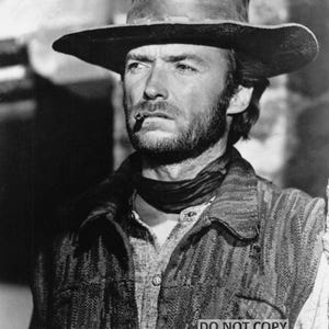 May include: Black and white image of a man wearing a cowboy hat, a vest, and a scarf. He has a beard and a cigarette in his mouth. The image is a portrait of a classic Western film character.