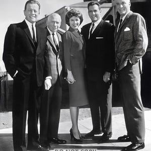 May include: Black and white photograph of five people, three men and two women, posing outdoors. The people are dressed in formal attire, including suits and dresses. The image has a vintage feel.