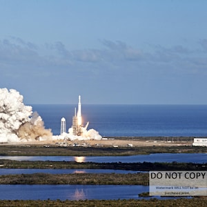 May include: A white rocket launches from a launchpad, leaving a trail of smoke and fire. The rocket is surrounded by a large cloud of smoke. The launchpad is located on a sandy beach with a body of water in the background. The sky is blue and clear.