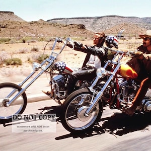 May include: Two men riding custom-built motorcycles on a desert road. The motorcycles are chrome and have a yellow and red paint job. The men are wearing leather jackets and jeans.