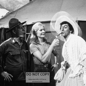 May include: Black and white photograph of three people. A person in a military uniform smokes a cigar. A woman lights a cigar for a person in a dress and a large hat. The scene appears to be outdoors, with a tent in the background.
