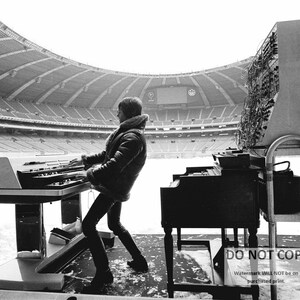 May include: Black and white photograph of a musician playing a keyboard and a piano on a stage in a stadium. The musician is wearing a jacket with a fur collar. The stadium is empty.