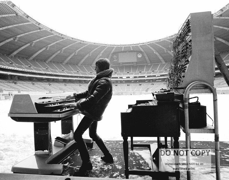May include: Black and white photograph of a musician playing a keyboard and a piano on a stage in a stadium. The musician is wearing a jacket with a fur collar. The stadium is empty.