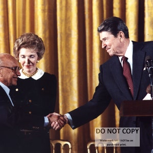 May include: A man in a suit shakes hands with another man in a suit. A woman in a black dress stands behind the man on the left. The background is a gold curtain.