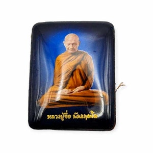 May include: A blue and gold framed image of a monk meditating. The image is set against a blue background and the monk is wearing a traditional orange robe. The text "หลวงพ่อ สมณะศิริ" is written in gold lettering at the bottom of the image.