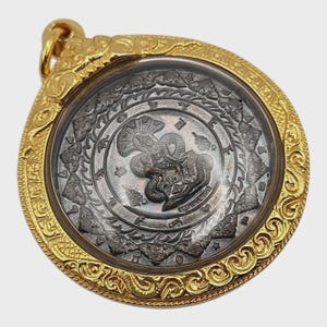 Könnte beinhalten: Goldfarbener Anhänger mit detailliertem, kunstvollem Design. Der Anhänger hat ein zentrales kreisförmiges Element mit einem detaillierten Bild einer Figur, umgeben von einem aufwendigen Rand. Der Anhänger hat eine Schlaufe zum Aufhängen.