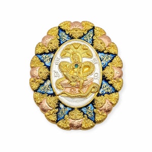 Könnte beinhalten: Ein Amulett aus Gold und Silber mit einem detaillierten Design einer Figur, die auf einem Thron sitzt, umgeben von einer Schlange. Das Amulett ist mit blauem und goldenem Email verziert und hat einen grünen Edelstein in der Mitte.