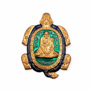 May include: A gold-colored turtle-shaped pendant with a central image of a seated monk. The pendant has a green and blue border with intricate detailing and text. The overall design is ornate and detailed.