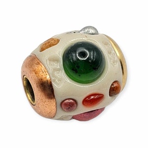 Peut inclure: Perle ovale blanche avec un œil en verre vert, entouré de plus petits accents en verre rouge, orange et brun. La perle est bordée de métal cuivre et or.