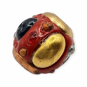 Könnte beinhalten: Eine handgefertigte Perle mit roter Basis, verziert mit goldenen, schwarzen und silbernen Metallic-Details. Die Perle hat eine unregelmäßige, abgerundete Form mit strukturierter Oberfläche. Das Design umfasst verschiedene Formen und Farben.