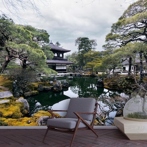 May include: A serene Japanese garden scene featuring a traditional pagoda reflected in a pond. Lush green trees and moss-covered rocks frame the water, with a modern gray chair and a glass terrarium in the foreground.