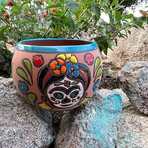 May include: A terracotta planter with a colorful Day of the Dead design featuring a woman's face with flowers in her hair. The planter has a turquoise rim.