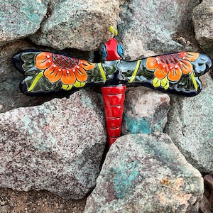 May include: A colorful ceramic dragonfly with red, orange, yellow, green, and blue paint. The dragonfly is decorated with floral patterns and is sitting on a bed of rocks.