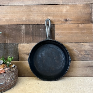 May include: A well-seasoned, black cast iron skillet with a long handle. The skillet is round with a slightly raised edge and a dark, textured cooking surface. The handle is silver-colored and has a hole for hanging. The background is a wooden wall.