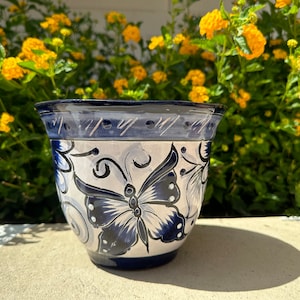 May include: A white ceramic planter with a blue rim and a blue butterfly design. The planter is sitting on a light gray surface.