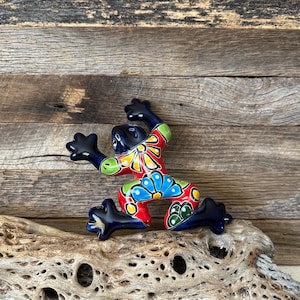 May include: A colorful ceramic frog figurine with a detailed floral design. The frog is painted with red, blue, yellow, and green accents. The frog is posed on a piece of driftwood against a rustic wooden background.
